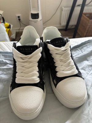 Svarta och vita sneakers från Numeris - Snygga sneakers från Numeris i svart tyg med vita detaljer och grova vita skosnören. Skorna har vit sula och tåparti i strukturmönstrat material. Låg modell med coola slitningar på sidan för en trendig look.