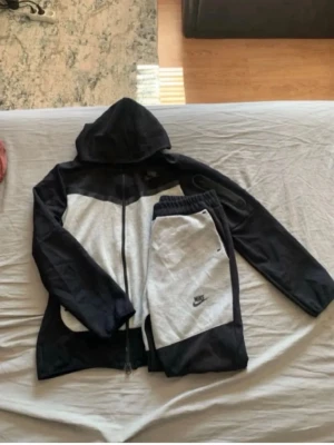 Nike svart och grått loungewear-set - Nike loungewear-set med zip-hoodie och matchande byxor i svart och ljusgrått. Hoodien har dragkedja och fickor, byxorna har resår i midjan och sidofickor. Båda plaggen har Nike-logga framtill. Perfekt för chill dagar hemma.