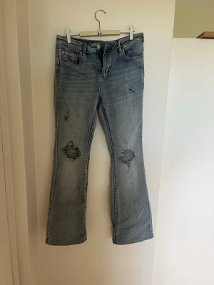 Ljusblå bootcut jeans - Säljer ett par ljusblå bootcut jeans med slitningar och hål på knäna. Jeansen är tillverkade i denim med en lätt tvättad look. Perfekta för en avslappnad och trendig stil.