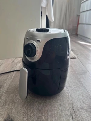 Svart airfryer med silverdetaljer - En kompakt airfryer i svart plast med silverfärgade detaljer och vred för temperatur och timer. Utrustad med handtag framtill och sladd för elanslutning. Perfekt för att laga mat utan olja.