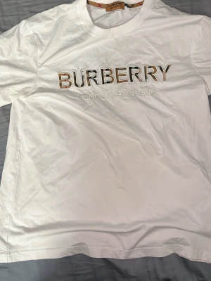 Vit Burberry t-shirt med logga - Vit t-shirt från Burberry med stor logga i beige, svart och vit på bröstet samt texten 'London England' under. Klassisk rund halsringning och korta ärmar. Tillverkad i mjuk bomull för en skön känsla och stilren look. Pris kan diskuteras vid snabb affär.