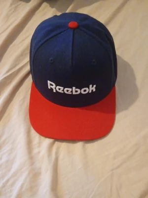 Blå och röd keps från Reebok - Snygg keps från Reebok i mörkblått tyg med röd skärm och röd knapp på toppen. Framtill finns Reebok-loggan broderad i vitt. Klassisk snapback-modell med justerbar passform och detaljerad insida med Reebok-band.