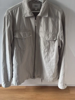 Ljusgrå overshirt från Adaher - Stilren ljusgrå overshirt med rak passform och två bröstfickor med lock. Dragkedja framtill och klassisk krage ger en clean look. Tillverkad i ett mjukt, strukturerat bomullstyg som känns bekvämt och passar till många outfits.