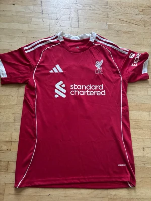 Liverpool matchtröja Adidas röd - Röd Liverpool FC matchtröja från Adidas med vita detaljer och tryck. Tröjan har klubbmärke, sponsorlogga och Adidas-logga på framsidan samt spelarnamn Wirtz och nummer 7 på ryggen. Tillverkad i lätt och ventilerande material, perfekt för fotbollsträning eller match.