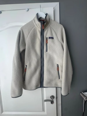 Beige fleecejacka från Patagonia - Säljer en beige fleecejacka från Patagonia med hög krage och hel dragkedja framtill. Jackan har tre fickor med dragkedja och snygga detaljer i brunt läder. Perfekt för kyliga dagar och riktigt skön att ha på sig.