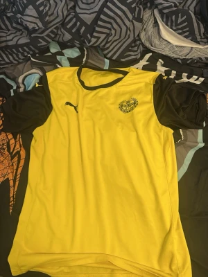 Gul och svart Puma fotbollströja - Snygg gul fotbollströja från Puma med svarta ärmar och detaljer. Tröjan har rund halsringning med svart kant och klubbmärke på bröstet. Perfekt för träning eller match och tillverkad i ett lätt och ventilerande material.