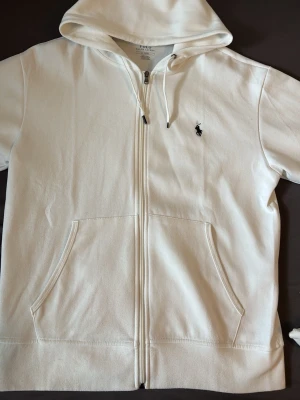 Vit zip hoodie från Polo Ralph Lauren - Snygg vit hoodie med dragkedja från Polo Ralph Lauren. Klassisk design med huva och dragsnören, två fickor framtill och den ikoniska lilla svarta logotypen broderad på bröstet. Perfekt för en clean och stilren look.