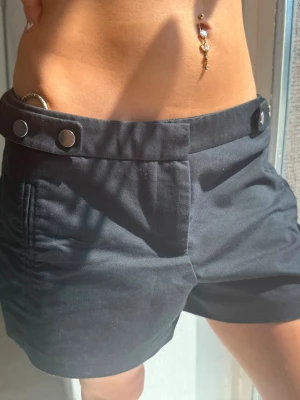 Svarta lågmidjade shorts med knappar - Snygga svarta shorts med låg midja och två silverfärgade tryckknappar framtill. Klassisk passform med fickor på sidorna och en bakficka. Perfekta för varma dagar och enkel att matcha med olika toppar.