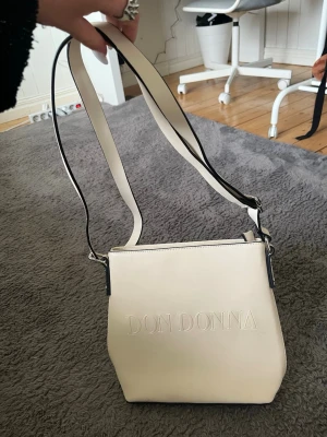 Beige axelväska från Don Donna - Snygg beige axelväska från Don Donna i stilren design. Väskan har justerbart axelband, präglad logga framtill och en praktisk dragkedjeficka på baksidan. Tillverkad i slätt konstläder med svarta detaljer längs kanterna.