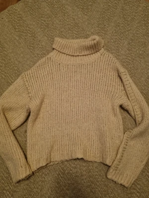 Beige stickad polotröja - Mysig beige stickad polotröja med grovstickat mönster och långa ärmar. Tröjan har ribbstickade muddar och en hög polokrage. Med guld detaljer 