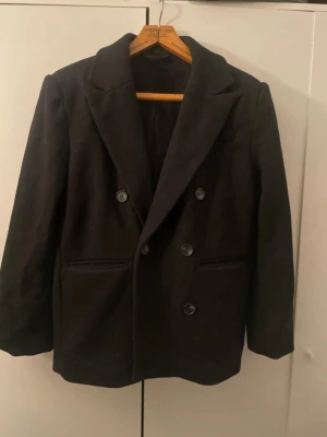 Svart dubbelknäppt kavaj Gina Tricot - Stilren svart kavaj från Gina Tricot i dubbelknäppt modell. Klassisk passform med breda slag och två framfickor. Tillverkad i ett slätt tyg med mjukt foder för extra komfort. Perfekt för en trendig och snygg look.