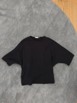 Svart loose topp med knyt bak - Svart topp i loose passform med korta ärmar och rund hals. Baksidan har en snygg knyt-detalj som ger extra stilpoäng. Perfekt för dig som gillar en enkel men trendig look.