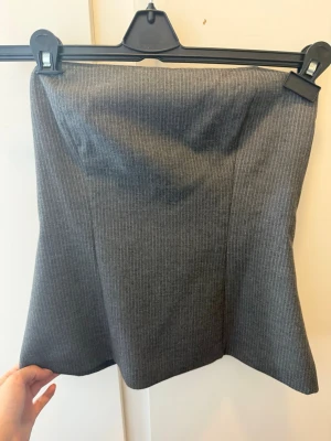 Grå randig tubtopp från Zara - Grå tubtopp från Zara med tunna vita ränder. Toppen har en enkel, rak passform och är axelbandslös, vilket ger en clean och stilren look. Perfekt att styla med höga jeans eller kjol.