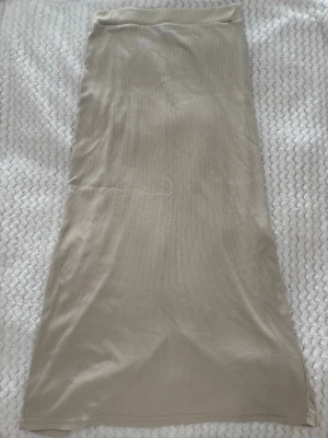 Beige ribbad långkjol med slits - Säljer en beige ribbad långkjol med slits bak. Kjolen har en enkel och stilren design med mjukt stretchigt material som sitter skönt på kroppen. Perfekt att matcha med en crop top eller hoodie för en chill look.