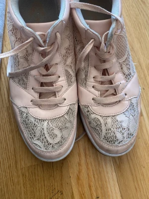 Rosa sneakers med spetsdetaljer - Unika sneakers i ljusrosa med snygga spetsdetaljer och matchande skosnören. Skorna har en vit sula och coola blommönster i spetsen som ger en trendig look. Perfekta för dig som vill sticka ut med din stil.