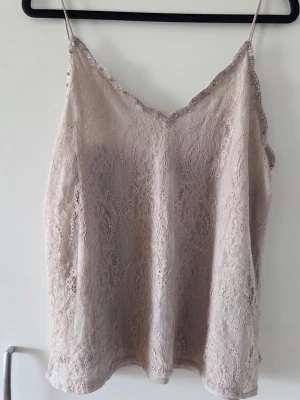 Beige spetslinne med tunna band - Säljer en ljusbeige linnetopp med tunna axelband och v-ringad hals som pryds av vackra strassdetaljer. Toppen är täckt av vacker spets och har en mjuk, feminin look. Perfekt för sommaren och enkel att styla med olika outfits.