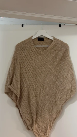 Beige stickad poncho med kabelmönster - Säljer en beige stickad poncho med snyggt kabelstickat mönster. Ponchon har en lös och avslappnad passform med rund halsringning. Perfekt att slänga över en tröja för extra värme. Materialet känns mjukt och bekvämt.
