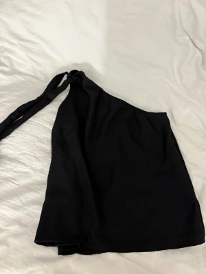 Svart oneshoulder topp med knyt - Säljer en svart oneshoulder topp med knyt i axeln. Toppen har en enkel och stilren design som passar perfekt till många olika outfits. Materialet känns mjukt och lätt, vilket gör den skön att bära under varma dagar.