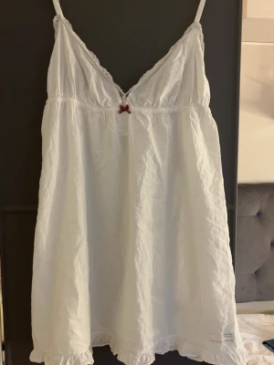 Vit klänning/ nattlinne med spets och rosett - Sött vitt klänning/ nattlinne med tunna axelband, spetskantad v-ringning och liten röd rosett framtill. Volangkant nertill och broderad patch med texten 'Lost on Purpose' på ryggen. Perfekt för mysiga kvällar hemma.