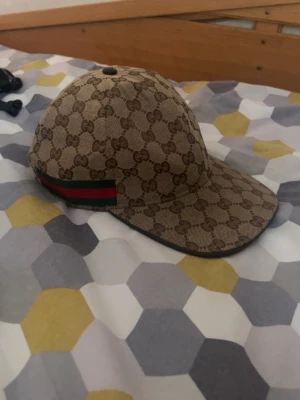 Gucci keps - Snygg beige keps från Gucci med klassiskt GG-monogram över hela kepsen och en grön-röd rand på sidan. Kepsen har böjd skärm och justerbar rem bak. Tillverkad i canvasmaterial med mörk insida och etikett på insidan.