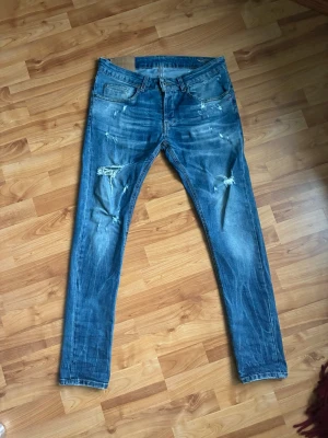 Blå Dondup jeans med slitningar - Säljer ett par blå Dondup George jeans med slitningar och hål på benen. Jeansen har klassisk femficksdesign, låg midja och är tillverkade i denim med stretch. Perfekta för en avslappnad och trendig look. W33 42cm i midjan och 102cm långa från midjan 