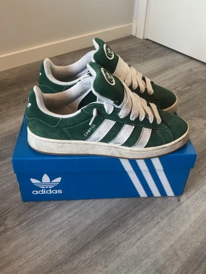 Adidas Campus 00s - Gröna Adidas Campus sneakers i mocka med vita detaljer och klassiska tre ränder på sidorna. Skorna har vit sula, breda vita skosnören och Adidas logga på plösen. Snygg retrostil som funkar till många outfits. Hör av er vid frågor, allt gott! 