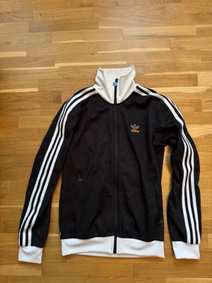 Svart Adidas track jacket med vita detaljer - Svart track jacket från Adidas med klassiska vita ränder längs ärmarna, vit krage, muddar och nederkant. Dragkedja framtill och två fickor med dragkedja. Adidas logga broderad på bröstet. Perfekt för en sportig och avslappnad stil.
