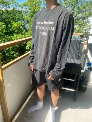 Svart långärmad topp Acne Studios - Cool svart långärmad topp från Acne Studios med vit text 'Acne Studios STOCKHOLM 1996' på bröstet. Oversized passform och rund halsringning. Perfekt för en avslappnad streetwear-look.