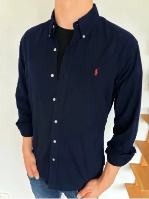 Mörkblå skjorta från Polo Ralph Lauren - Klassisk mörkblå skjorta från Polo Ralph Lauren med knappar framtill och röd broderad logga på bröstet. Skjortan har lång ärm, button-down krage och är tillverkad i bomull. Perfekt för en stilren och avslappnad look.