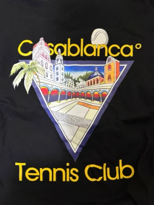 Svart Casablanca Tennis Club t-shirt - Snygg svart t-shirt från Casablanca med färgglatt tryck framtill som visar en tennisbana och byggnader, samt texten 'Casablanca Tennis Club' i gult. Klassisk passform och rund hals. Perfekt för dig som gillar streetwear och grafiska prints.