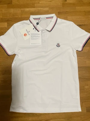 Vit Moncler pikétröja med röd detalj - Klassisk vit pikétröja från Moncler med röd och marinblå rand på krage och ärmslut. Tröjan har knappslå med tre knappar och Moncler-logga broderad på bröstet. Perfekt för en clean och stilren look.