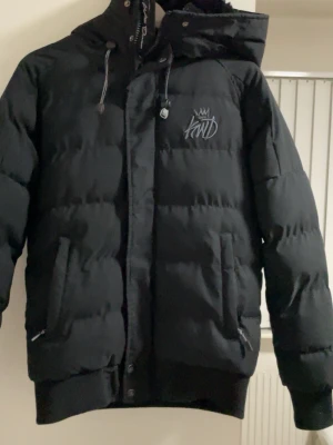 Svart pufferjacka från KWD - Säljer en svart pufferjacka från KWD med broderad logga på bröstet. Jackan har huva med dragsko, två framfickor med dragkedja och ribbade muddar vid ärmslut och nederkant. Perfekt för kalla dagar och riktigt snygg streetstil.
