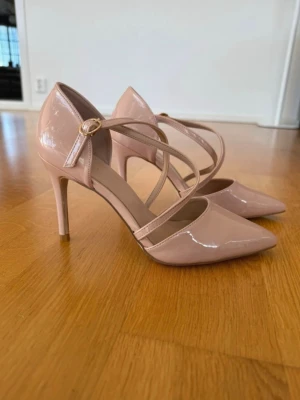 Nude lackpumps med remmar - Säljer ett par snygga pumps i nude/rosa färg med glansig lackyta och spetsig tå. Skorna har smala korsade remmar med guldfärgat spänne och hög klack. Perfekta för dig som vill ha en elegant och stilren look.
