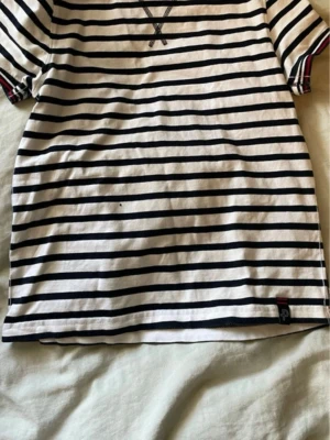 Randig t-shirt från Pelle P - Säljer en klassisk randig t-shirt från Pelle P i vitt och marinblått. T-shirten har korta ärmar, rund hals med röd och marinblå kant och är tillverkad i mjuk bomull. Perfekt för en avslappnad och stilren look.