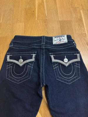 Mörkblå bootcut jeans True Religion - Säljer ett par mörkblå bootcut jeans från True Religion med vita sömmar och fickor med blingdetaljer. Midjemått: 34cm.🩷 Innerbenslängd: 76cm.🩷