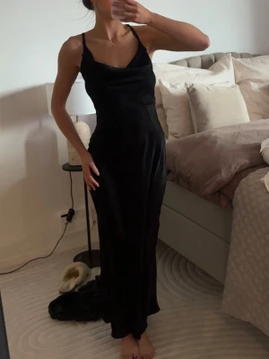 Svart långklänning - Säljer en elegant svart långklänning med smala axelband och öppen rygg. Klänningen har en mjuk följsam passform och lätt glansigt material som ger en lyxig känsla. Perfekt för dig som vill ha en stilren och tidlös look.