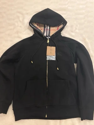 Svart hoodie från Burberry - Snygg svart hoodie från Burberry med dragkedja och klassiskt rutigt foder i huvan. Guldiga detaljer på snörena och diskret broderad logga på bröstet. Tillverkad i mjuk bomull för maximal komfort. Perfekt för en stilren och avslappnad look.