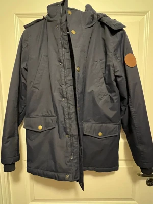 Mörkblå parkas med huva - Snygg mörkblå parkas med avtagbar huva och stora fickor framtill. Jackan har tryckknappar i mässingsfärg och en rund läderpatch på ärmen. Materialet är slitstarkt och vattenavvisande, perfekt för kalla dagar. Broderad logga och siffran 27 på ryggen.