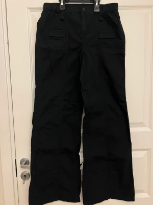 Svarta cargobyxor med vida ben - Säljer ett par svarta cargopants med breda ben och elastisk midja. Byxorna har stora fickor framtill med lock och bälteshällor. Tillverkade i ett slitstarkt bomullsmaterial och har en loose fit som ger en avslappnad look. Aldrig använda.