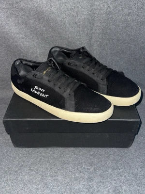 Svarta Saint Laurent sneakers - Svarta sneakers från Saint Laurent med vit logga broderad på sidan. Skorna har svart canvas och mockadetaljer, samt vita gummisulor. Klassisk låg modell med svarta skosnören och diskret branding på plösen. Perfekta för en clean och stilren look.