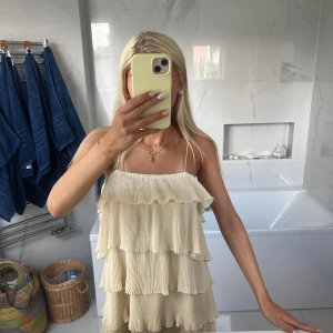 Plisserad volangklänning i ljusbeige - Supersöt kort klänning med tunna axelband och flera lager plisserade volanger. Ljusbeige färg som ger en fräsch och somrig vibe. Perfekt för varma dagar och snygg att styla med smycken. Materialet känns lätt och luftigt.