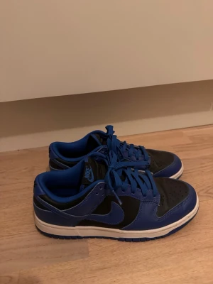 Nike Dunk Low blå och svarta sneakers - Säljer ett par Nike Dunk Low sneakers i blått och svart med vit sula. Skorna har klassisk låg profil, perforerad tå och Nike-logga på sidan. Materialet är en mix av läder och textil, perfekt för dig som vill ha en stilren och sportig look.
