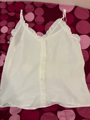 Vit linnetopp med volangkant - Söt vit linnetopp med tunna axelband och volangdetaljer längs v-ringningen och framtill. Perfekt för varma dagar och enkel att styla med jeans eller kjol. Materialet känns lätt och luftigt.