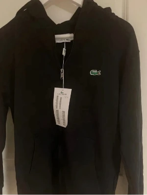 Svart hoodie från Lacoste med dragkedja - Svart hoodie från Lacoste med klassisk krokodillogga på bröstet. Modellen har dragkedja framtill, huva och känguruficka. Tillverkad i mjukt bomullsmaterial som är skönt att bära. Perfekt för en avslappnad och stilren look.