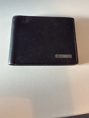 Svart läderplånbok från ben sherman  - Snygg svart plånbok i äkta läder från ben Sherman’s Klassisk fyrkantig modell med flera kortfack och ett genomskinligt fack för ID eller körkort. Stilren design med metallogo på framsidan. Perfekt för dig som vill ha en enkel och praktisk plånbok. 