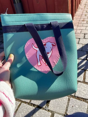 Turkos shoppingväska med hundmotiv - Turkos shoppingväska från Ulla Martelius i konstläder med mörkblå axelband. Framsidan har ett rosa hjärta med en tecknad hund i vitt och rött. Väskan har en enkel, fyrkantig form och är perfekt för att bära med sig det viktigaste.