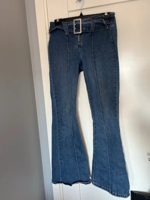 Blå utsvängda jeans med bälte - Säljer ett par blå jeans med utsvängda ben och markerade sömmar framtill och baktill. Jeansen har ett brett bälte med stor silverfärgad spänne i midjan. Klassisk denimlook i mjukt material, perfekta för en trendig och avslappnad stil.