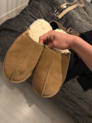 Beige fårskinnstofflor med ullfoder - Mysiga beige tofflor i mocka med fluffigt vitt ullfoder. Klassisk slip-in modell som håller fötterna varma under kalla dagar. Perfekta för att chilla hemma och ge en skön känsla. Tidlös design utan synligt märke.