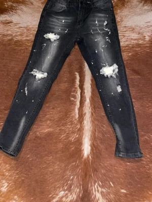 Svarta skinny jeans med slitningar - Säljer ett par svarta skinny jeans med coola slitningar och blekta detaljer på benen. Jeansen har klassisk femficksmodell och är i stretchigt denimtyg. Perfekta för en trendig streetstyle-look.