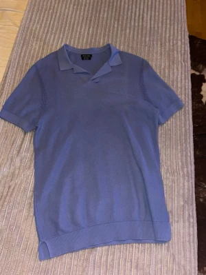 Ljusblå pikétröja från Massimo Dutti - Snygg ljusblå pikétröja från Massimo Dutti i exklusiv bomull. Tröjan har korta ärmar, klassisk krage och ribbade muddar nedtill. Perfekt för en stilren och avslappnad look.